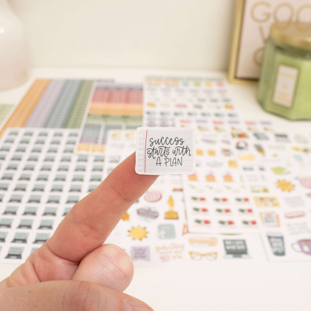 Hand holding a motivational planner sticker that says 'Success starts with a plan', with various planner stickers in the background.