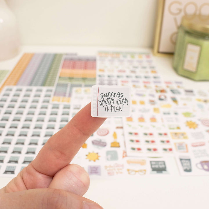 Hand holding a motivational planner sticker that says 'Success starts with a plan', with various planner stickers in the background.