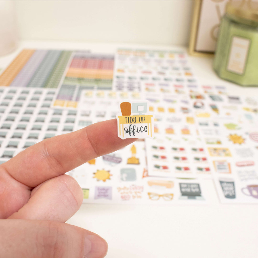 Close-up of a 'Tidy Up Office' sticker held on a finger, showcasing Bloom Workday Essentials Planner Stickers.