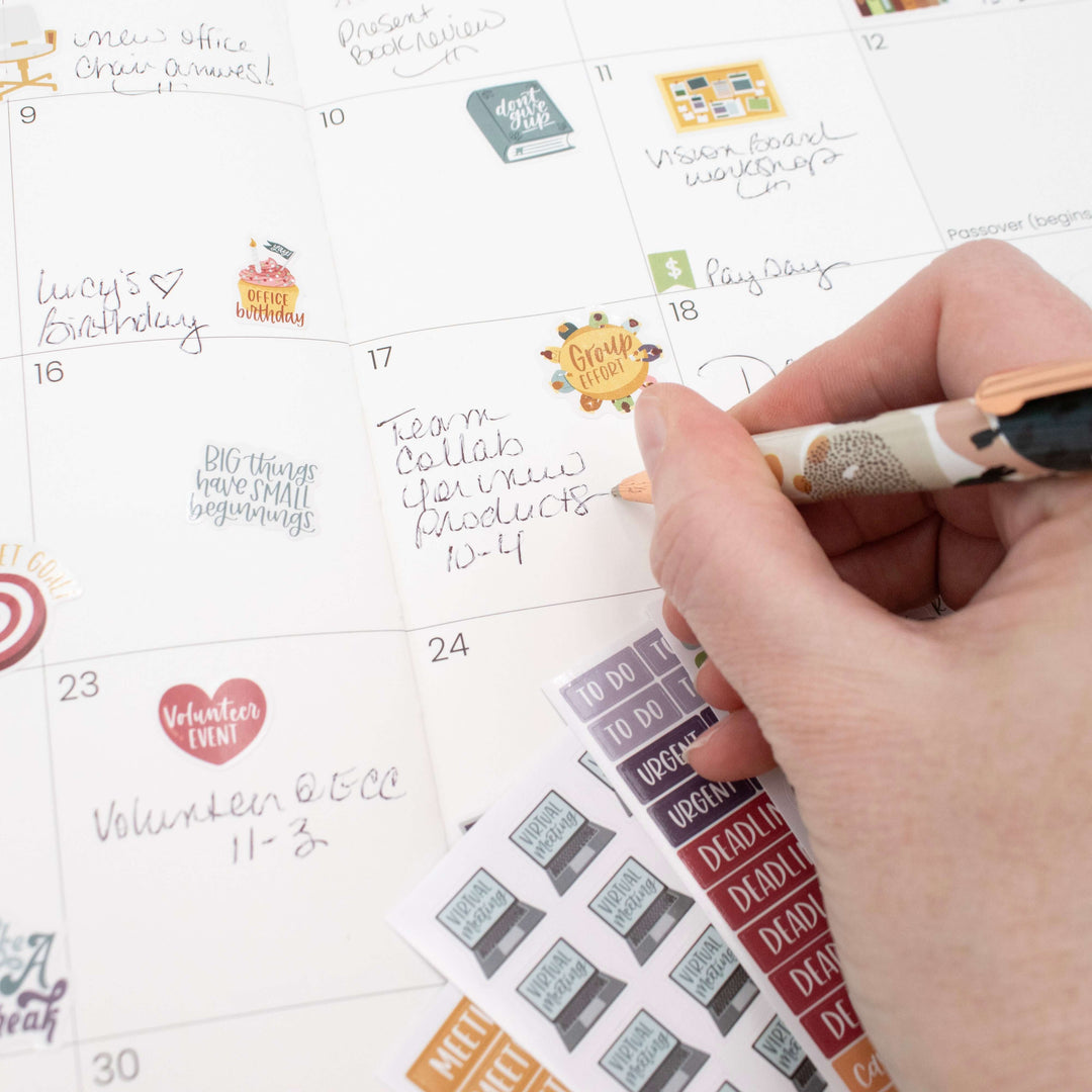 Hand using a pen on a calendar with Bloom Workday Essentials Planner Stickers for organization.