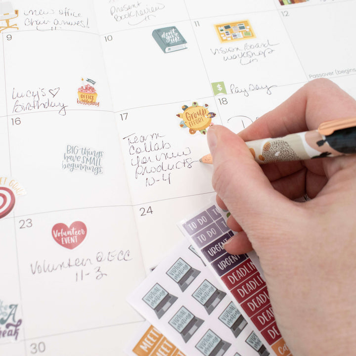 Hand using a pen on a calendar with Bloom Workday Essentials Planner Stickers for organization.