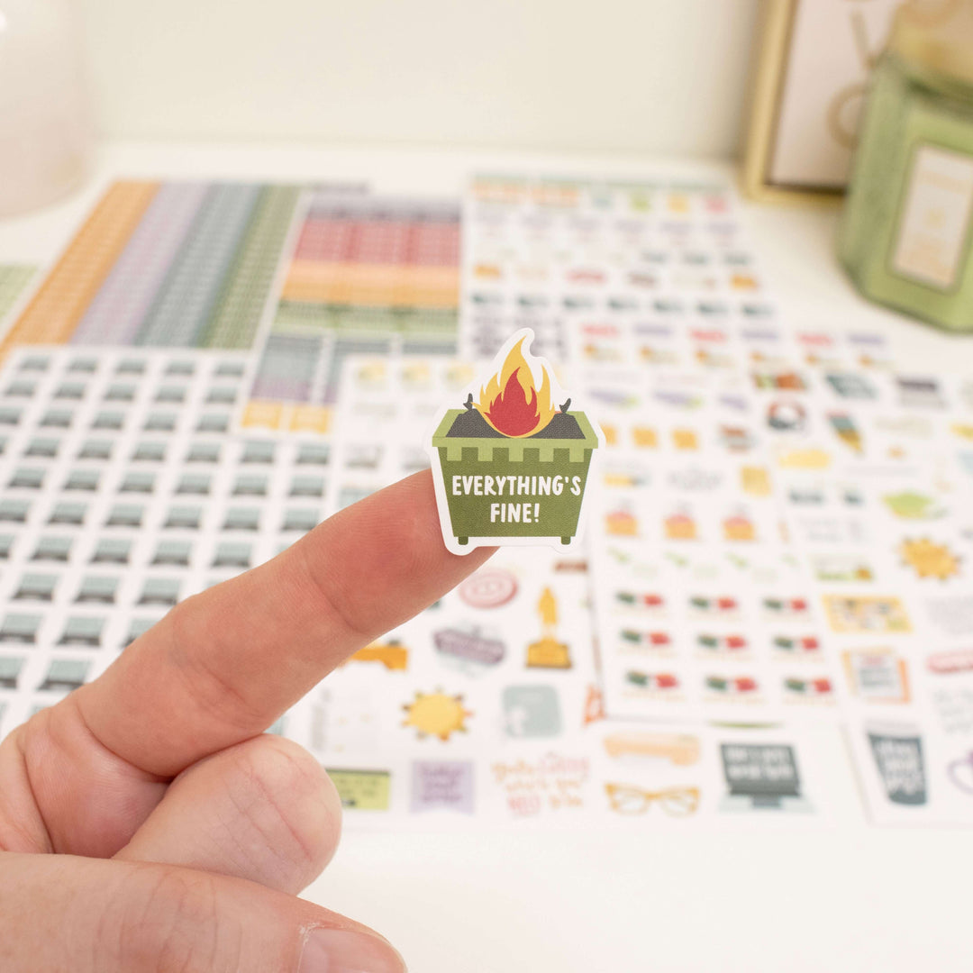 Hand holding a 'Everything's Fine!' planner sticker from Bloom Workday Essentials Sticker Pack with organized backgrounds.