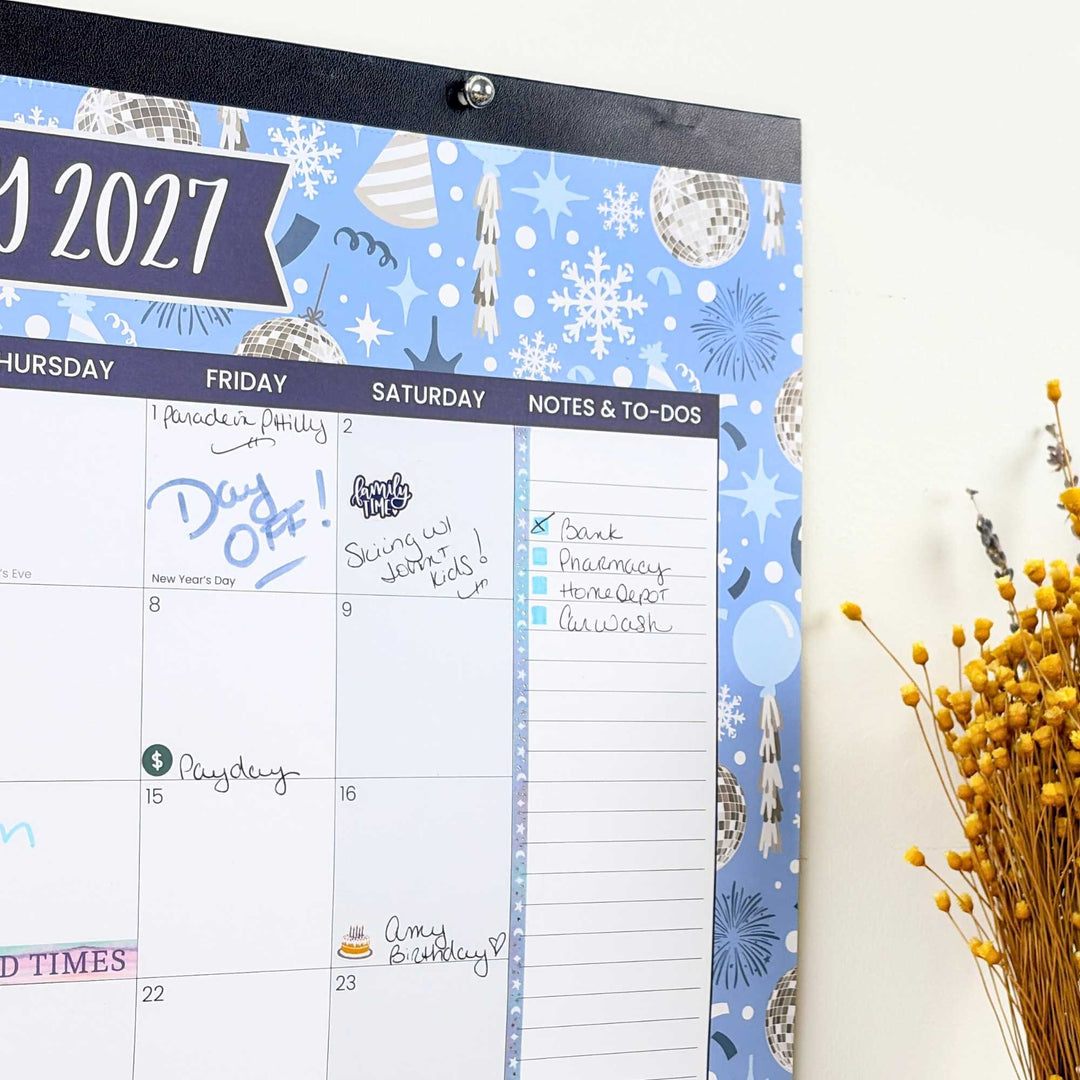 2027 calendar with decorative cover featuring snowflakes and party hats, next to a vase of dried flowers.