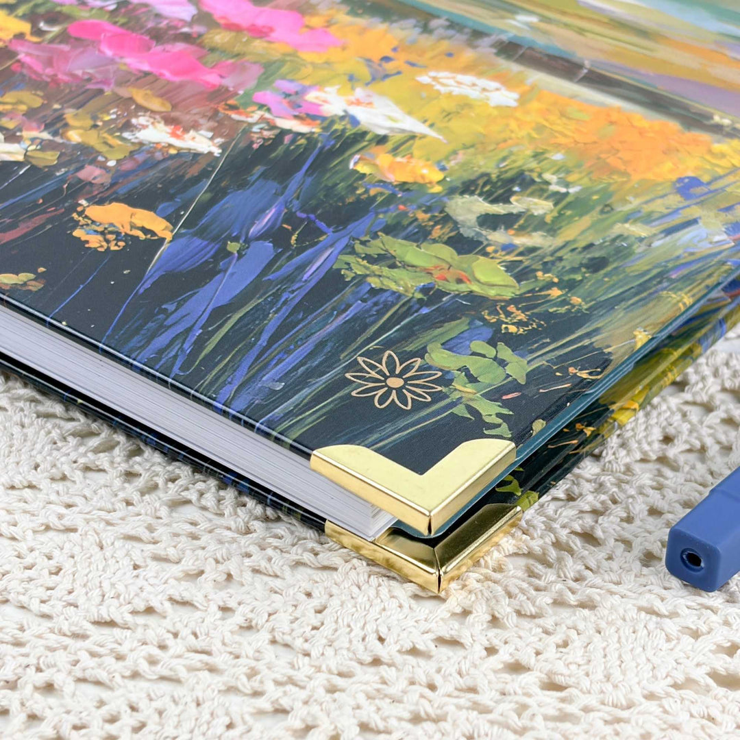 A hardcover notebook or journal with a colorful floral and landscape-style painted cover, shown resting on a cream knit blanket. The cover features a dark navy base with vivid blue, green, yellow, pink, and orange brushstrokes, plus gold corner protectors.