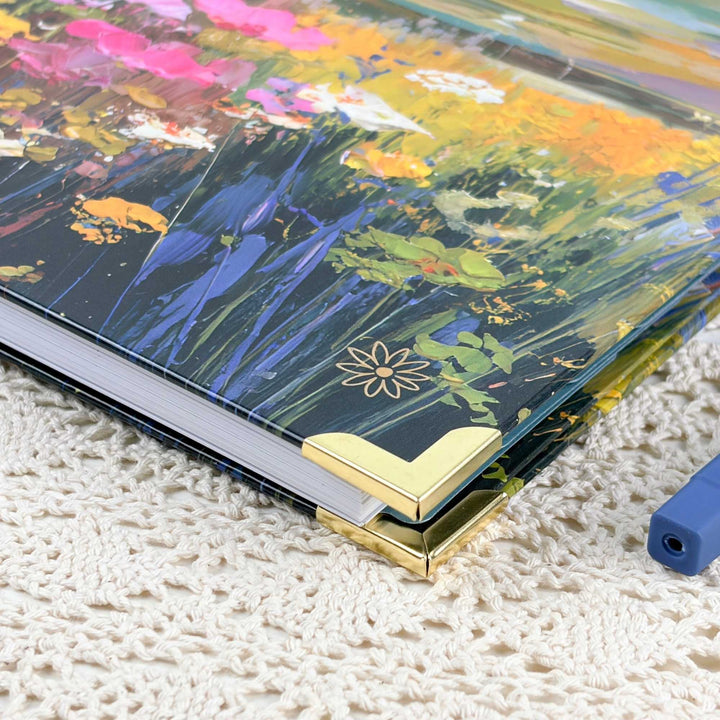A hardcover notebook or journal with a colorful floral and landscape-style painted cover, shown resting on a cream knit blanket. The cover features a dark navy base with vivid blue, green, yellow, pink, and orange brushstrokes, plus gold corner protectors.