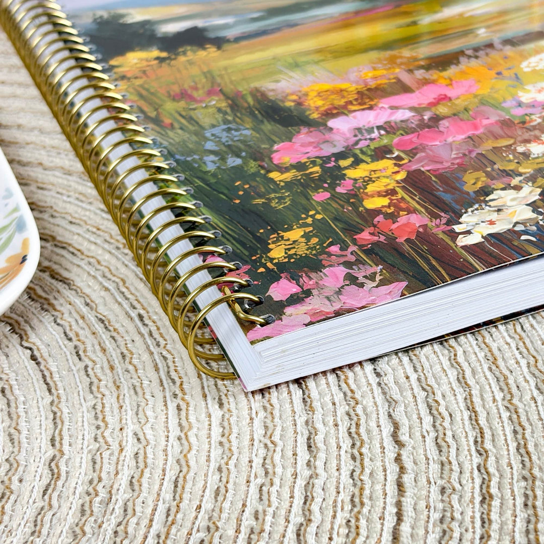 A spiral-bound notebook with a gold wire binding and a colorful floral landscape cover, resting on a textured beige woven placemat.