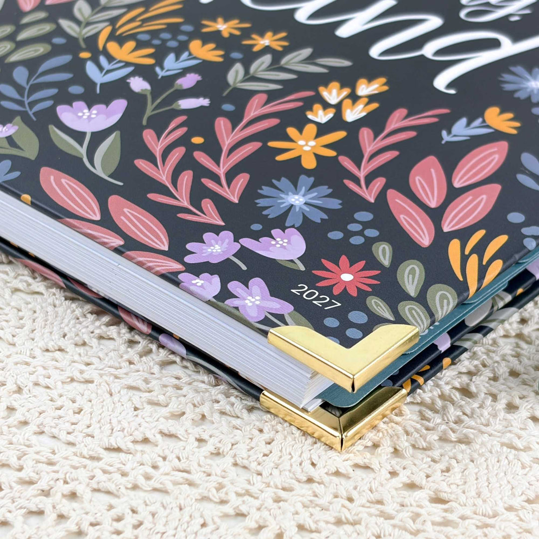 A close-up of a 2027 floral-bound book or journal with a dark cover covered in colorful spring flowers and leaves, resting on a cream-colored textured knit blanket. The book has gold metal corner protectors.