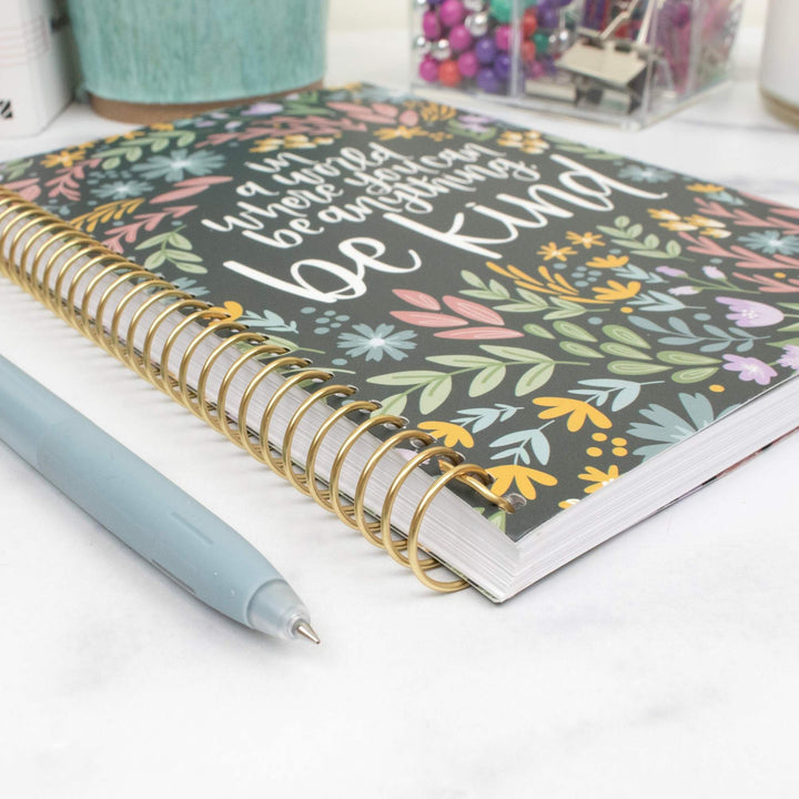 A spiral-bound notebook with a dark gray cover decorated with colorful floral illustrations and the phrase 'You are worth it because you are kind' in white script. A light blue pen lies across the foreground on a white marble-like surface, with office supplies blurred in the background.