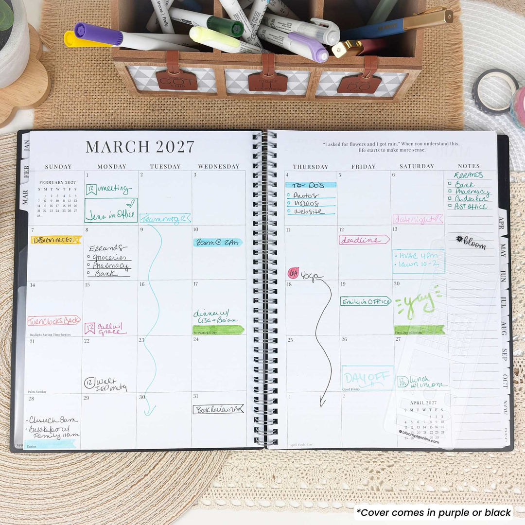 An open spiral-bound planner showing the monthly view for March 2027, laid flat on a woven mat with pens, clips, and other stationery around it. The planner pages are white with black grid lines and small colored notes, and a small caption at the bottom says the cover comes in purple or black.