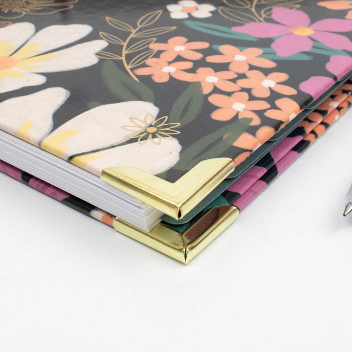 A close-up of a floral notebook or journal with metallic gold corner protectors, showing a dark gray cover decorated with pink, orange, and white flowers.