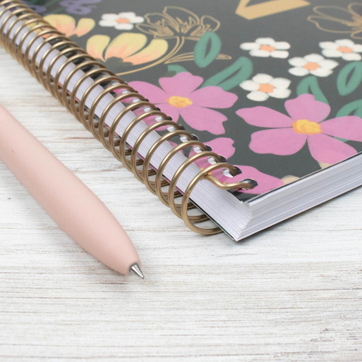 A spiral-bound notebook with a floral cover design in pink, yellow, white, and green on a dark background, shown on a light wood surface next to a pale pink pen.