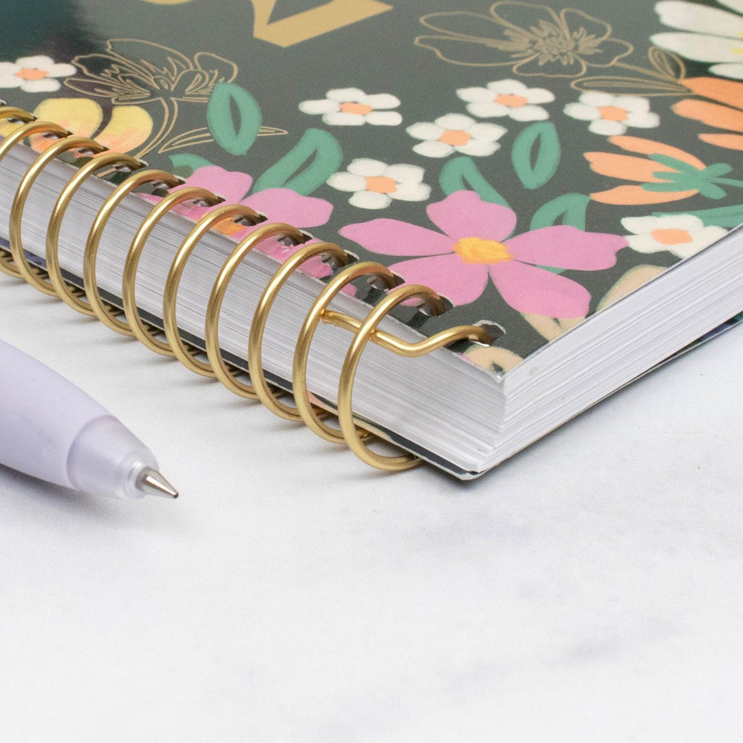 A close-up of a spiral-bound notebook with a gold wire coil and a floral cover, placed on a light marble-like surface next to a white pen.
