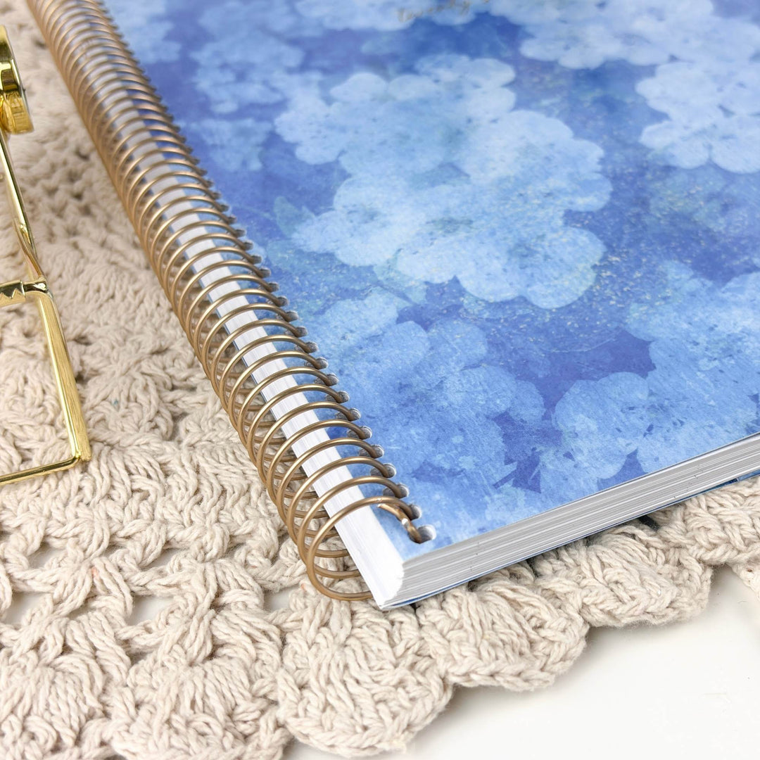 A spiral-bound notebook with a blue watercolor floral cover, resting on a beige crocheted surface next to a gold metal stand.