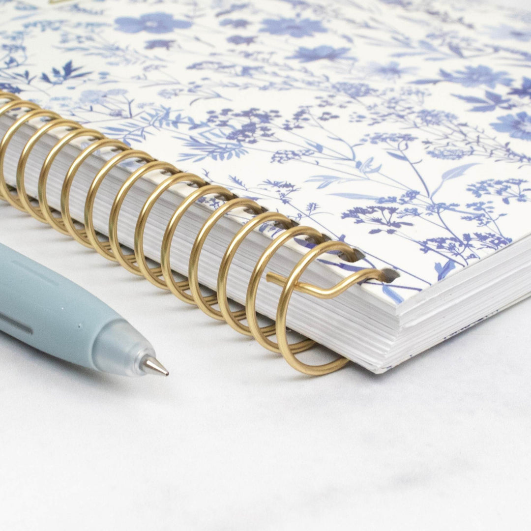 A close-up of a spiral-bound notebook with a blue floral pattern on a white cover and a gold wire spiral binding, shown on a light marble-like surface with a pale blue pen nearby.