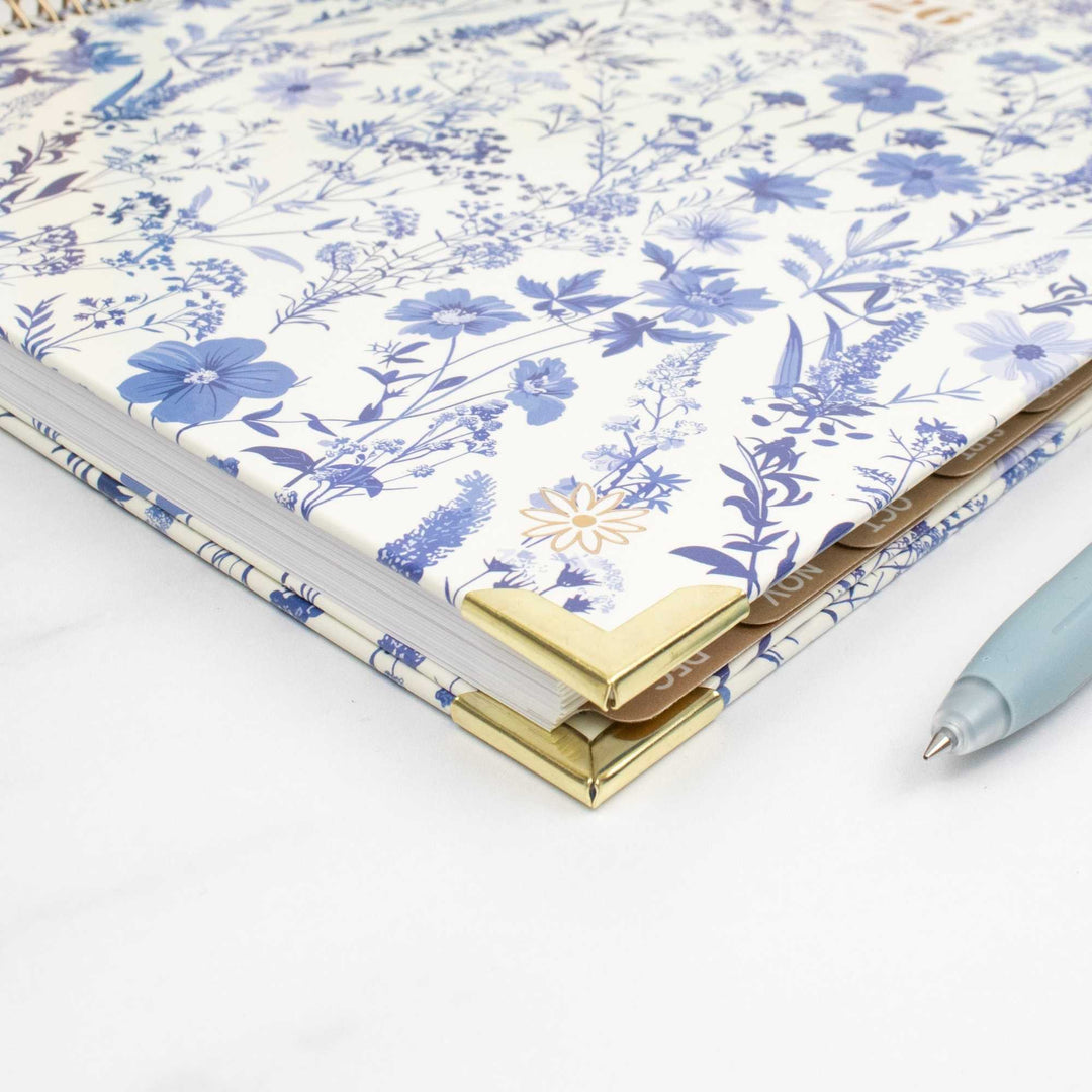 A close-up of a floral notebook or planner with a white cover decorated in blue flowers and green leaves, featuring gold metal corner protectors. A light blue pen is partially visible on the right side against a clean white background.