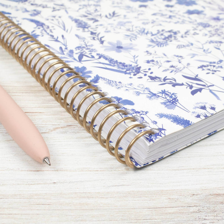 A spiral-bound notebook with a blue floral toile pattern on white paper, shown on a light wood surface next to a pale pink pen.