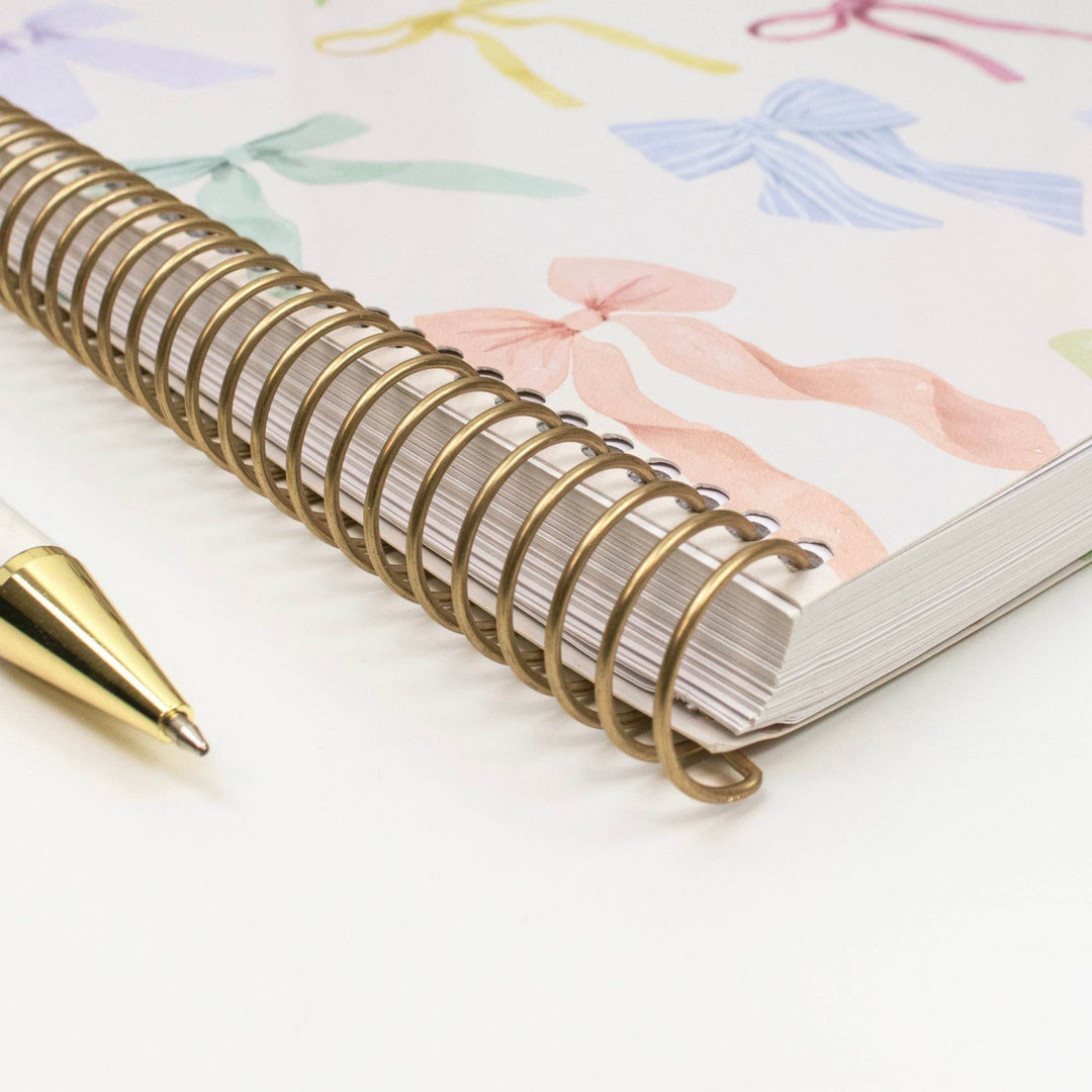 A close-up of a spiral-bound notebook with a gold wire coil and a white cover decorated with pastel bow illustrations, shown on a white surface with a gold pen nearby.