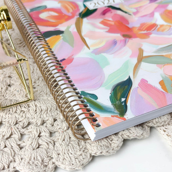 A spiral-bound 2027 planner with a colorful floral cover design, styled on a beige crocheted surface next to a gold geometric desk accessory.