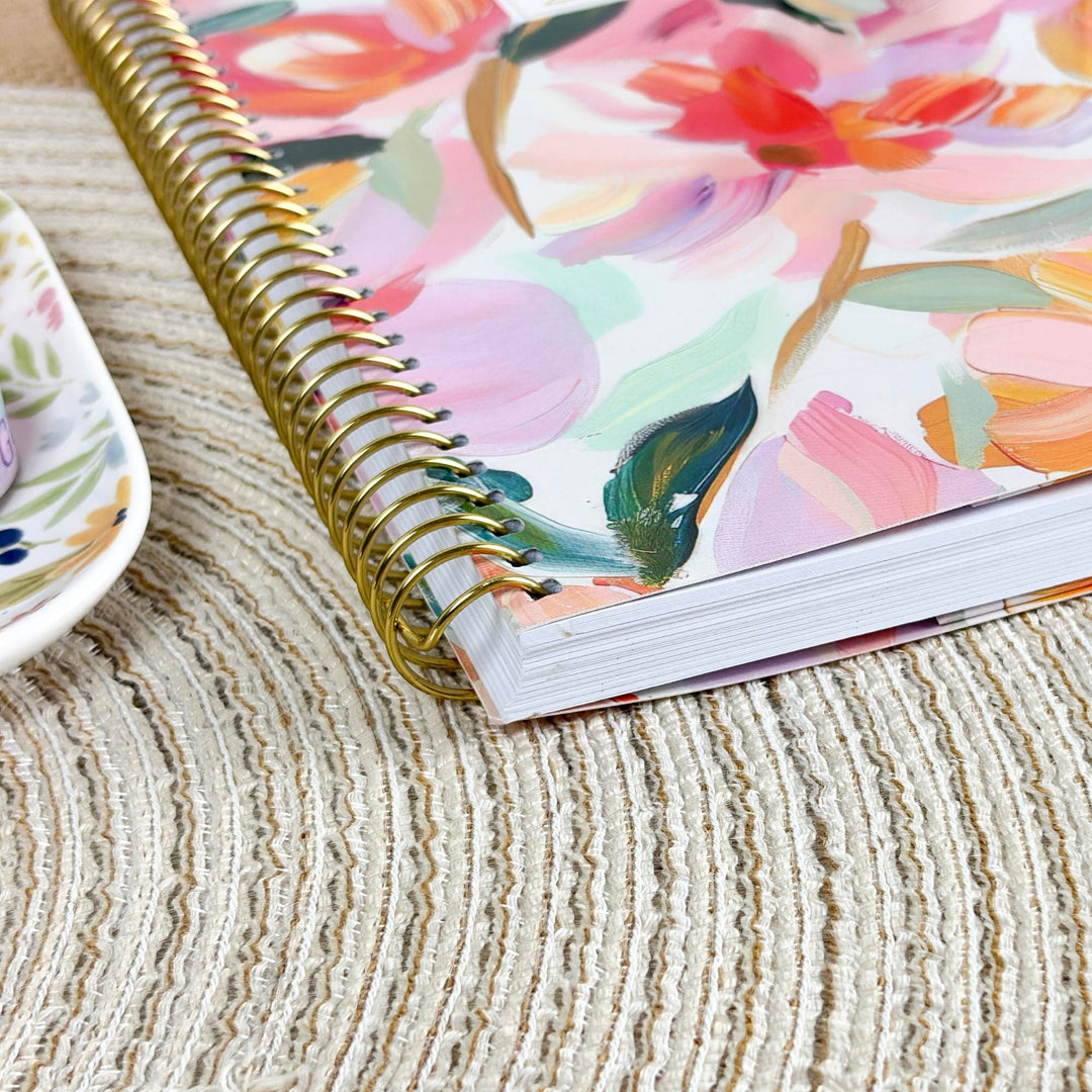 A close-up of a spiral-bound notebook with a colorful floral cover, resting on a textured beige woven placemat. A partial white plate with a floral pattern is visible on the left edge.