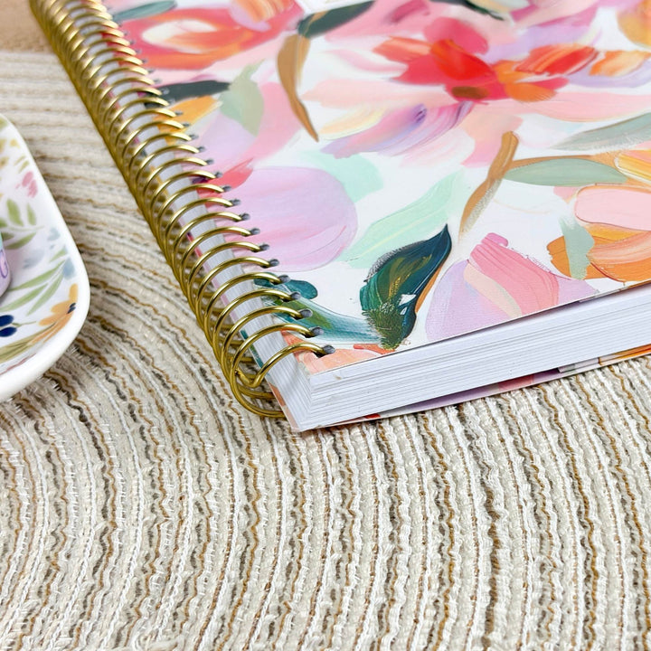 A close-up of a spiral-bound notebook with a colorful floral cover, resting on a textured beige woven placemat. A partial white plate with a floral pattern is visible on the left edge.