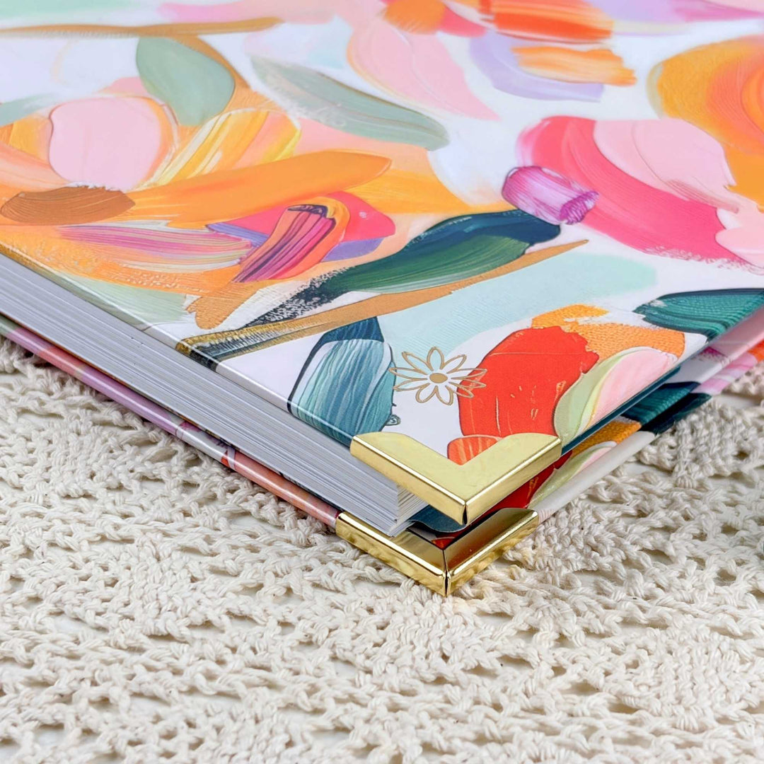 A close-up of a hardcover notebook or journal with a colorful floral cover design, resting on a cream-colored crocheted surface. The cover features a vibrant, painterly floral pattern in pink, orange, yellow, green, and blue, with a gold metal corner accent.