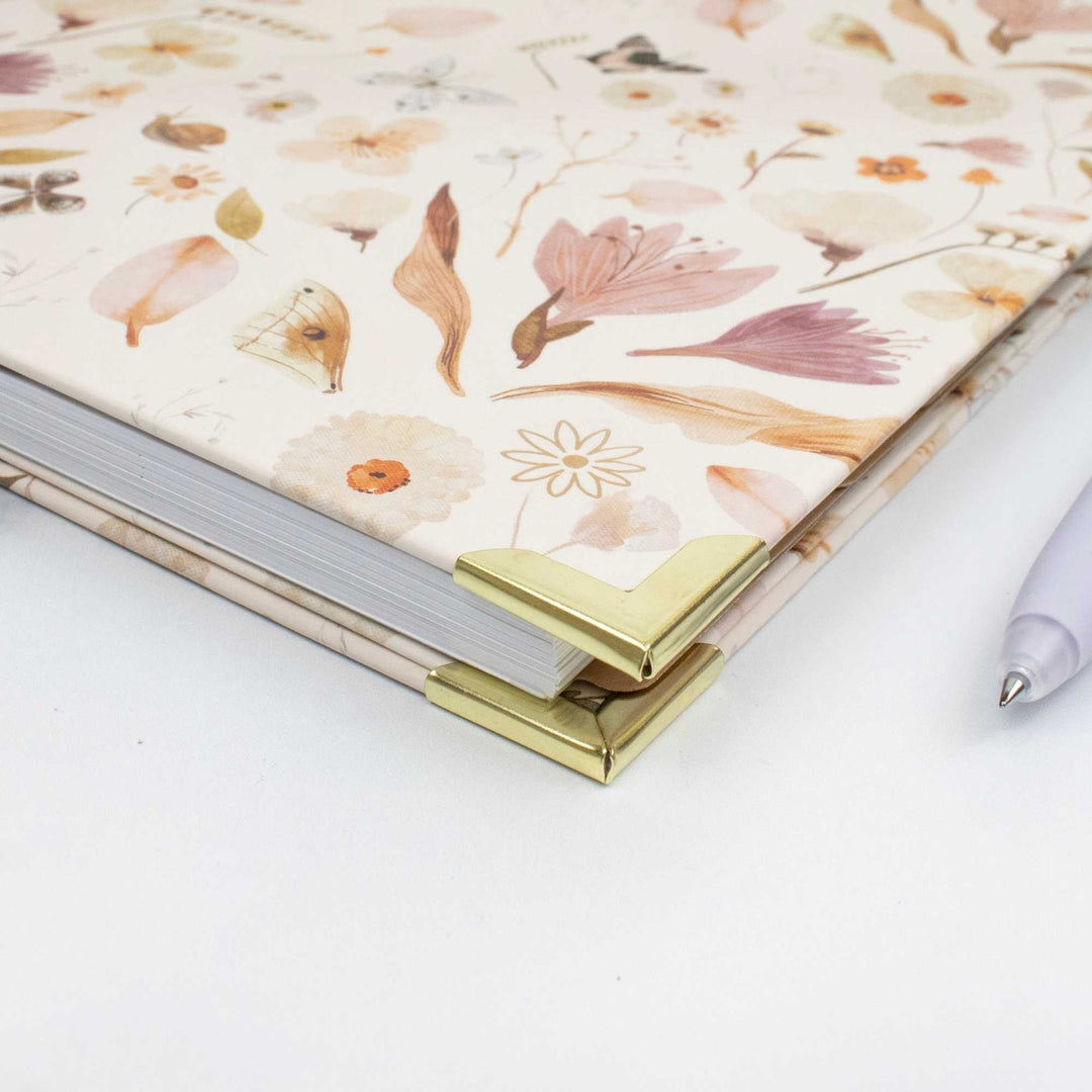 A hardcover notebook with a cream-colored cover decorated with a delicate pastel floral and botanical pattern, featuring shiny gold metal corner protectors, shown on a white surface next to a light purple pen.