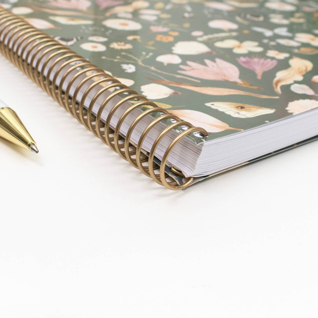 A close-up of a spiral-bound notebook with a muted green floral cover and a gold-tone wire spiral, placed on a white surface with part of a matching gold pen visible.