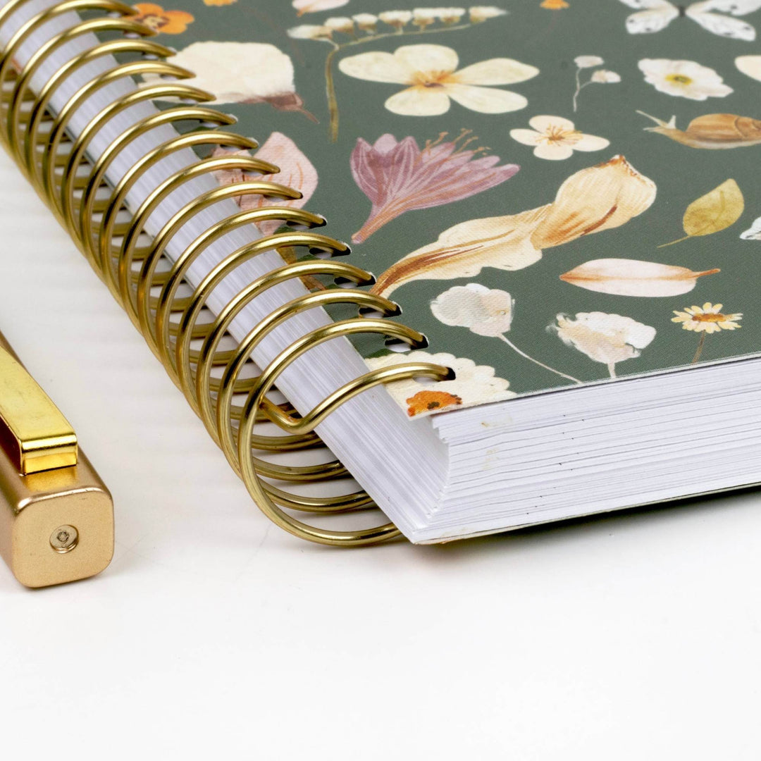 A close-up of a spiral-bound notebook with a muted green floral cover and a gold wire spiral, shown on a white surface with part of a gold pen visible at the lower left.