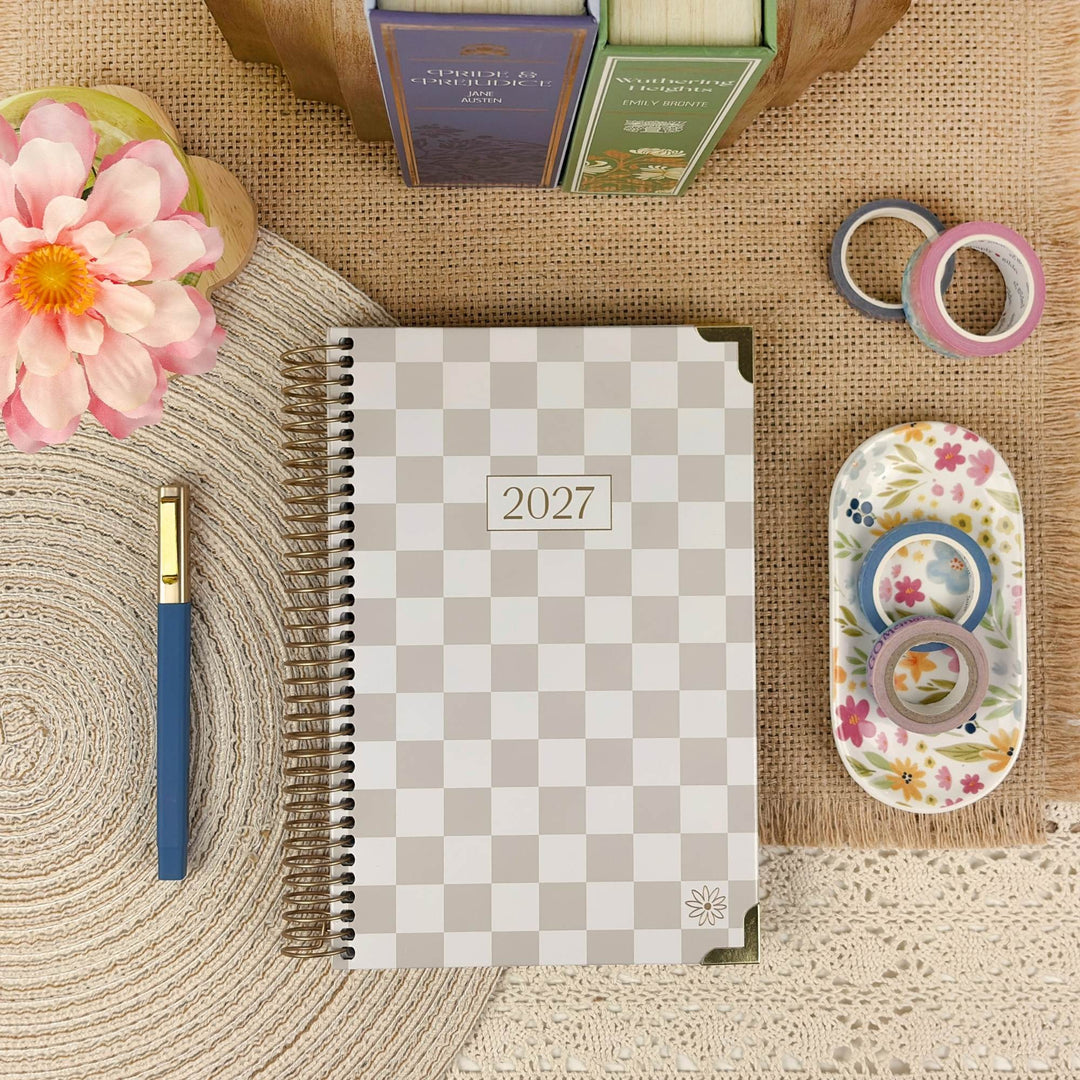 A 2027 spiral-bound planner with a beige and gray checkered cover is styled in a flat lay on a burlap surface, surrounded by a pink flower, a blue pen, washi tape rolls, and a floral dish.