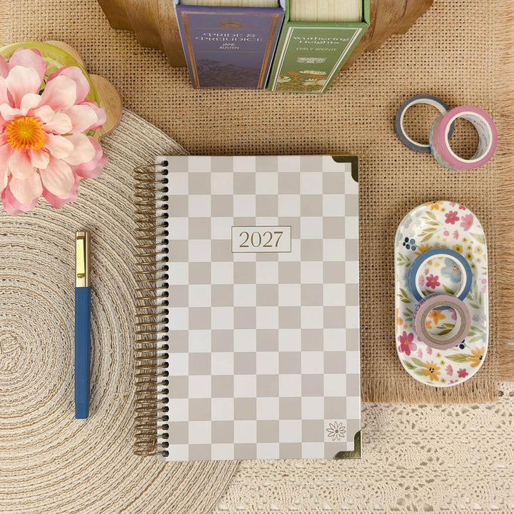 A 2027 spiral-bound planner with a beige and gray checkered cover is styled in a flat lay on a burlap surface, surrounded by a pink flower, a blue pen, washi tape rolls, and a floral dish.