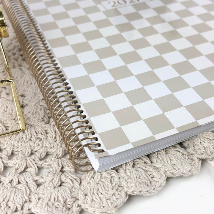 A spiral-bound 2027 planner with a beige and white checkerboard cover, resting on a textured cream crochet blanket next to a gold metal stand.