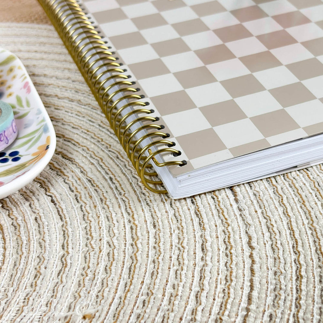 A spiral-bound planner or notebook with a beige and white checkerboard cover, shown open on a textured cream woven mat. A small floral dish is partially visible on the left.
