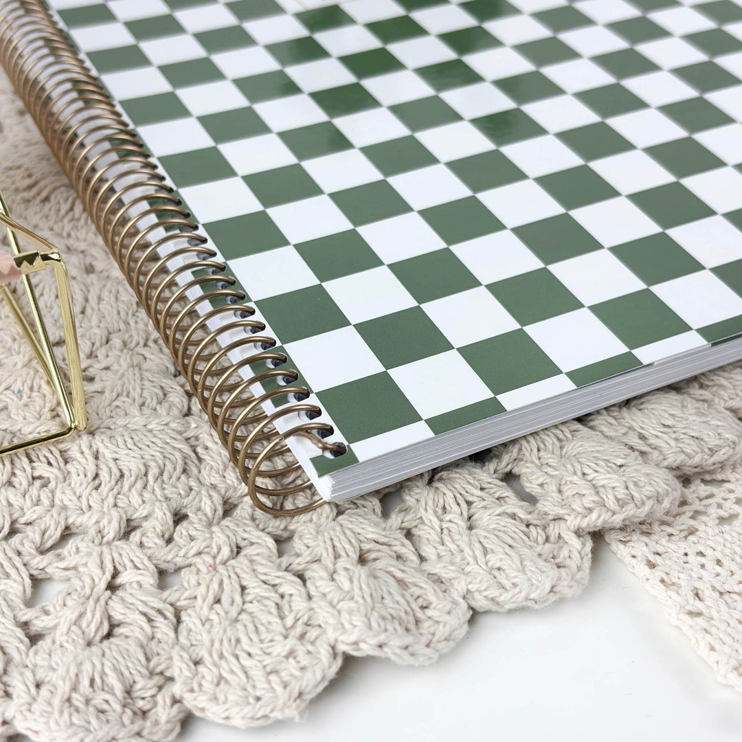 A spiral-bound notebook with a green and white checkerboard cover, styled on a beige crocheted mat next to a small gold geometric decor object.
