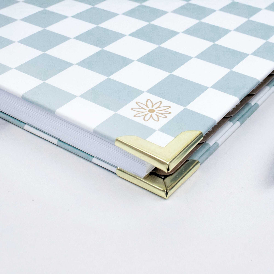A close-up of a hardcover notebook or journal with a light blue and white checkerboard pattern, featuring a gold foil flower design on the cover and gold metal corner protectors.