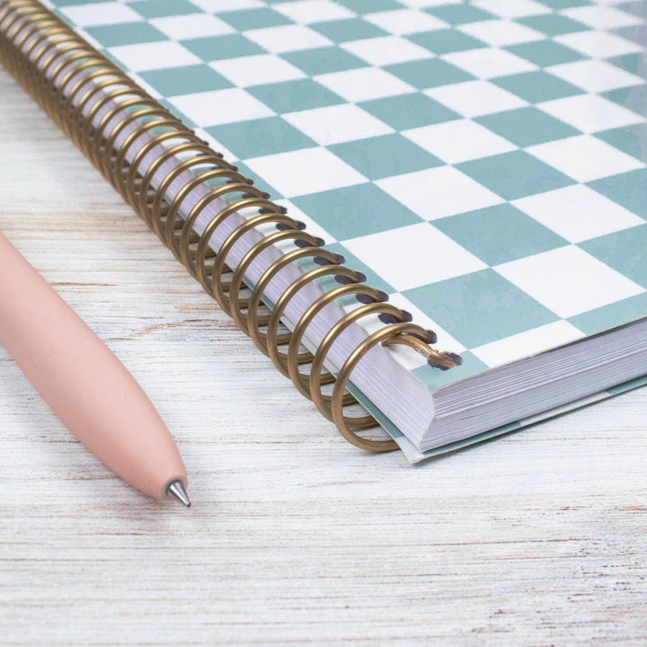 A spiral-bound notebook with a mint green and white checkerboard cover lies on a light wood surface next to a pale pink pen.