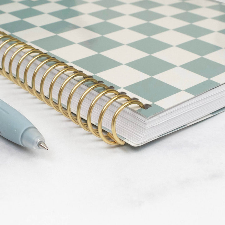 A spiral-bound notebook with a mint green and white checkerboard cover, bound with a shiny gold metal coil, shown on a white marble-like surface next to a light blue pen.