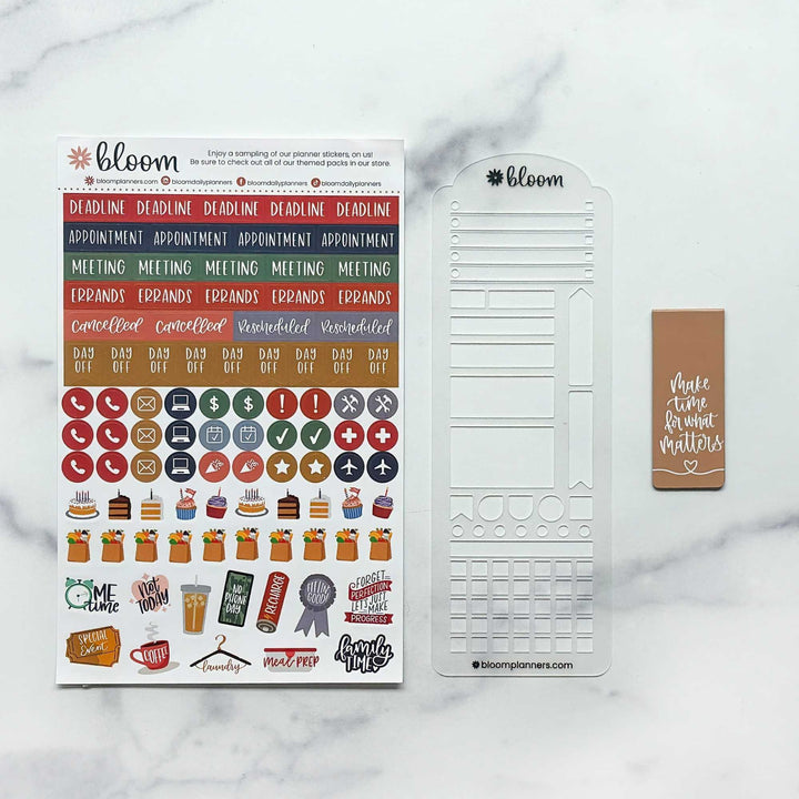 Bloom planner sticker kit displayed on a white marble background, featuring a sheet of colorful planner stickers, a white planner insert sheet, and a brown sticky note with white text.