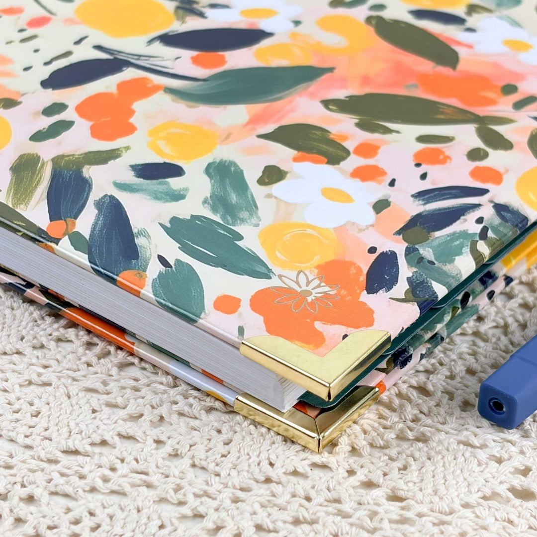 A close-up of a hardcover notebook or journal with a colorful floral and abstract paint-splatter cover, resting on a cream-colored crocheted surface. The cover features orange, yellow, green, and navy brushstrokes on a light background, with gold-toned metal corner protectors and white page edges visible.