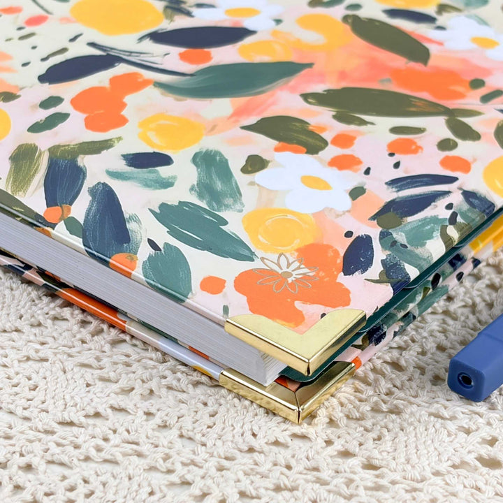 A close-up of a hardcover notebook or journal with a colorful floral and abstract paint-splatter cover, resting on a cream-colored crocheted surface. The cover features orange, yellow, green, and navy brushstrokes on a light background, with gold-toned metal corner protectors and white page edges visible.