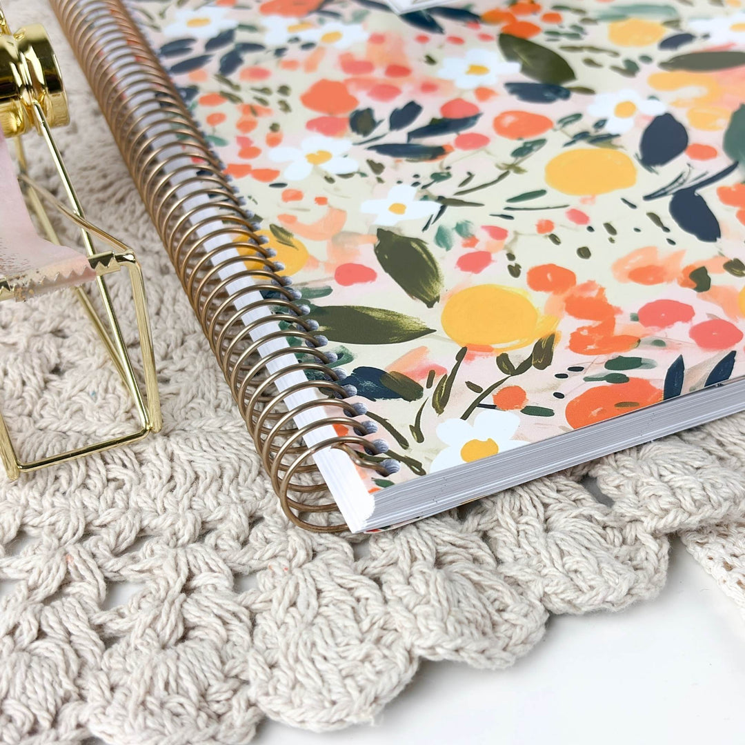 A spiral-bound notebook with a colorful floral cover lies on a cream crocheted surface next to a small gold-framed decorative object.