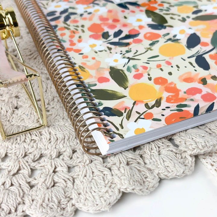 A spiral-bound notebook with a colorful floral cover lies on a cream crocheted surface next to a small gold-framed decorative object.