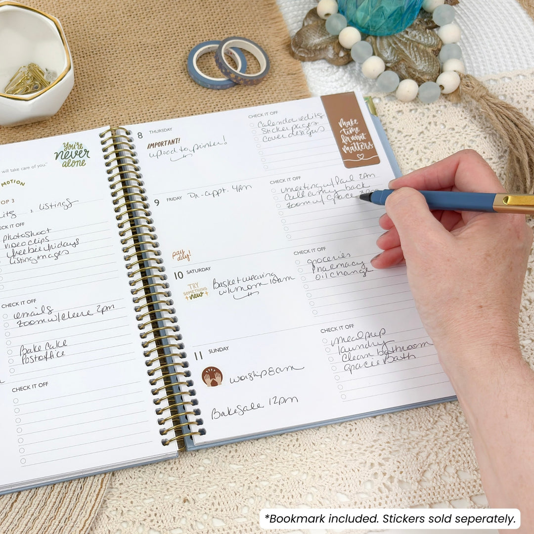 An open spiral-bound planner on a textured beige surface, with a hand holding a blue pen pointing to the page. The planner has white pages with handwritten notes, gold-toned spiral binding, and a small brown sticker that reads 'Plan Your Week.