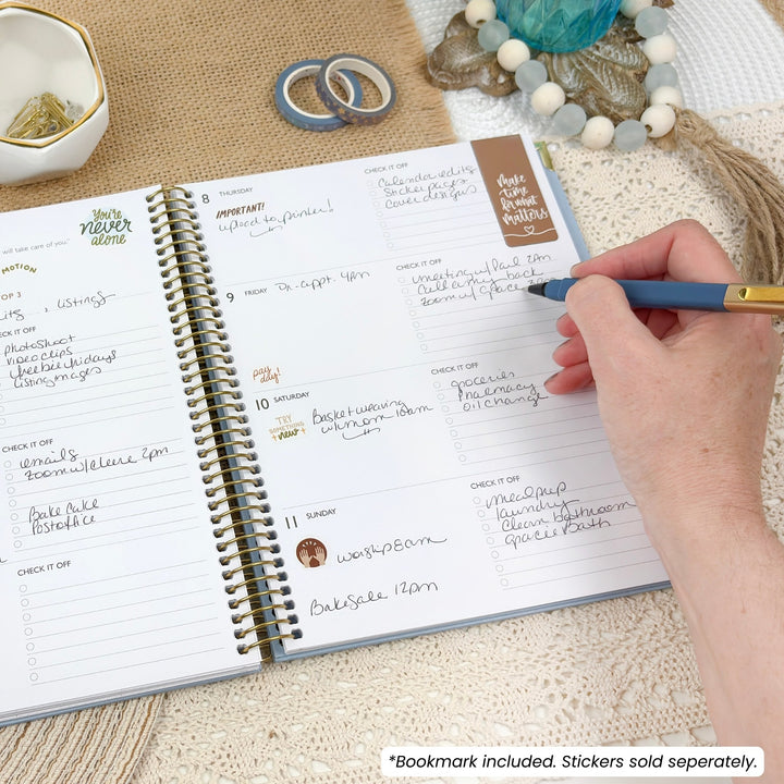 An open spiral-bound planner on a textured beige surface, with a hand holding a blue pen pointing to the page. The planner has white pages with handwritten notes, gold-toned spiral binding, and a small brown sticker that reads 'Plan Your Week.