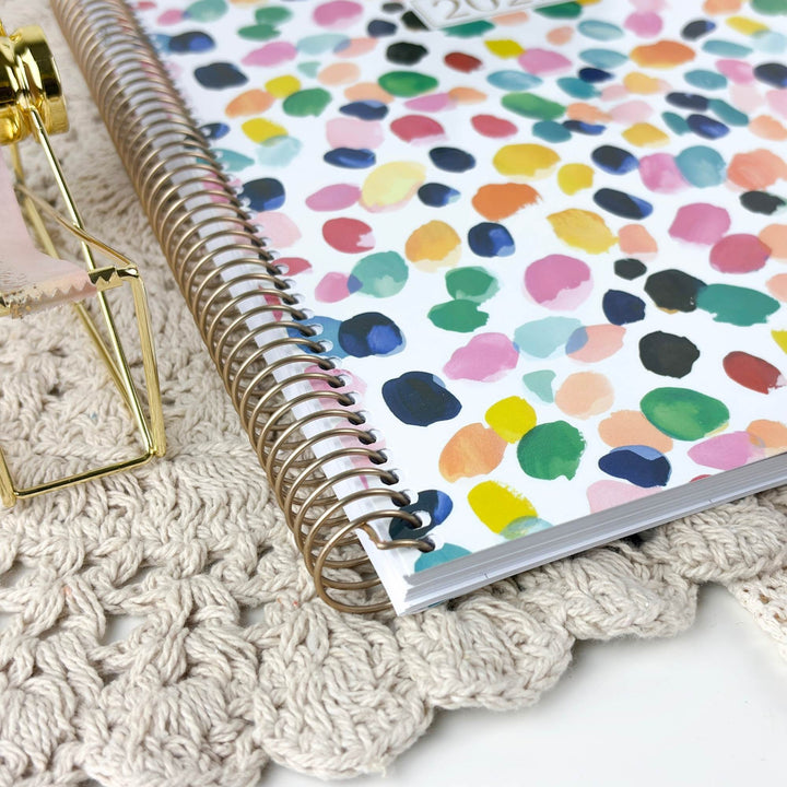 A spiral-bound notebook with a white cover decorated with colorful watercolor polka dots, resting on a beige crocheted rug next to a gold-framed decorative object.