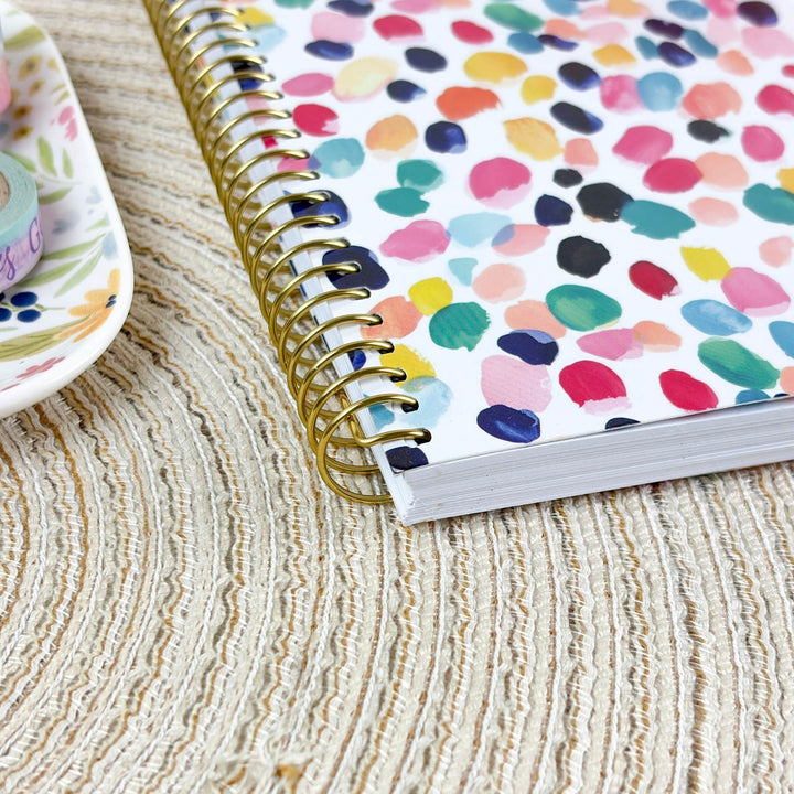 A spiral-bound notebook with a white cover covered in colorful watercolor paint dots, resting on a beige woven placemat next to a floral dish.