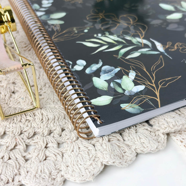 A spiral-bound notebook with a black cover decorated with gold botanical line art and watercolor eucalyptus leaves, styled on a cream-colored crocheted mat next to a gold-framed object.