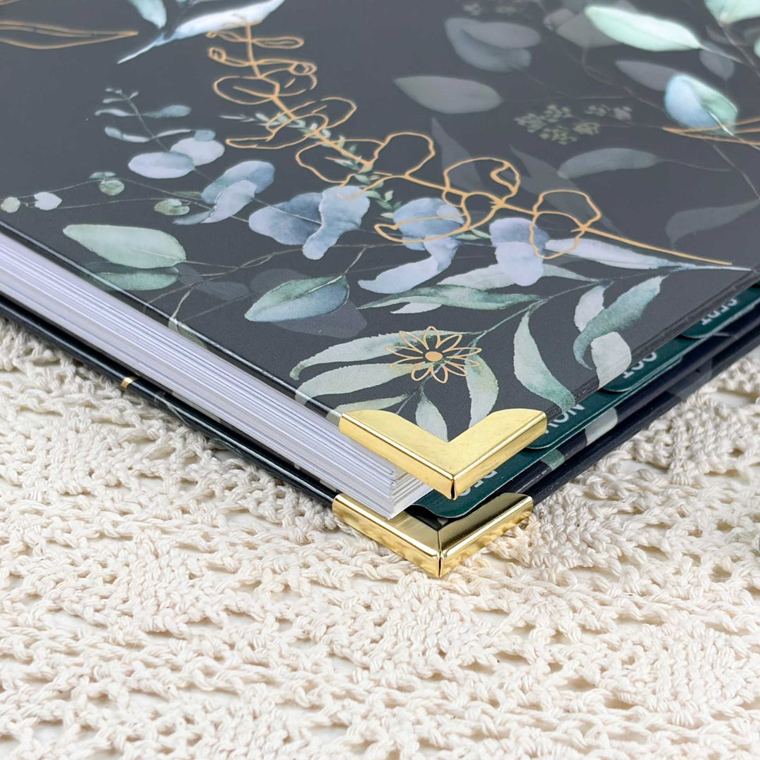 A close-up of a hardcover notebook or journal with a dark gray floral cover featuring eucalyptus leaves and gold foil details, resting on a cream-colored textured lace surface.