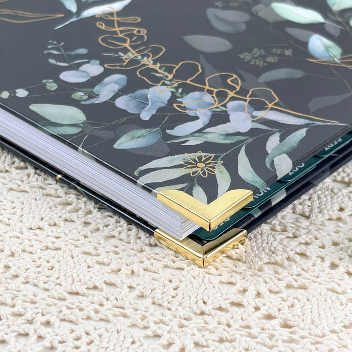 A close-up of a hardcover notebook or journal with a dark gray floral cover featuring eucalyptus leaves and gold foil details, resting on a cream-colored textured lace surface.