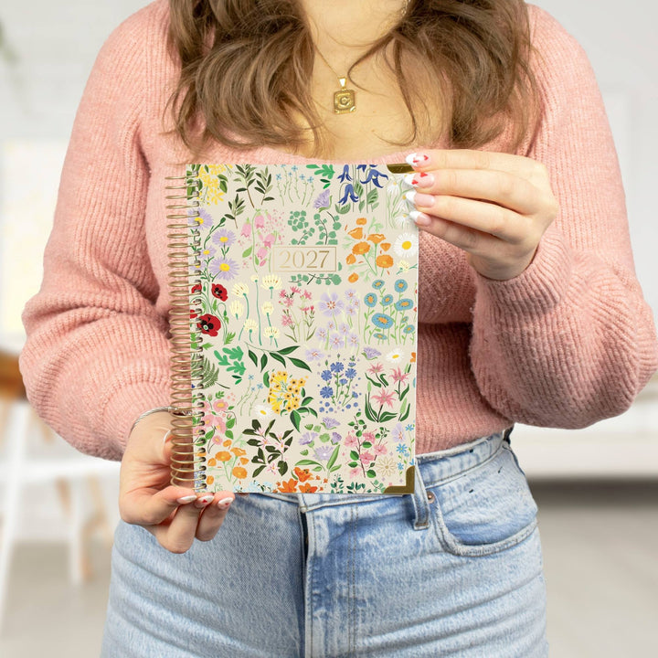 A 2027 spiral-bound planner with a floral cover design, shown with its monthly divider pages and a white sticker sheet branded bloom.
