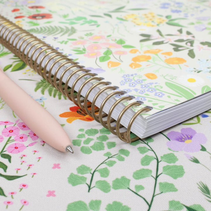 A floral-patterned spiral notebook with a gold wire coil binding, shown on top of a matching floral fabric surface with a pale pink pen resting beside it.